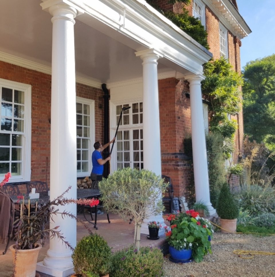 Andrew cleaning tall sash windows behind the columns of a large period building